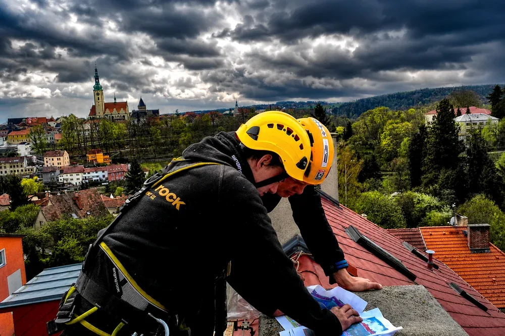 Výškový specialista Výškové práce Anděl v přilbě a lanovém postroji studuje plány na střeše v Táboře, v pozadí panorama města s věží kostela – obhlídka a příprava pracovního postupu horolezeckou technikou